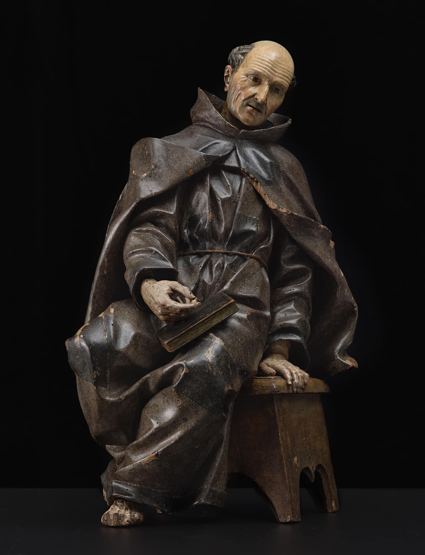 a wood sculpture of a man dressed in a monk's costume sitting on a bench with a book in his lap