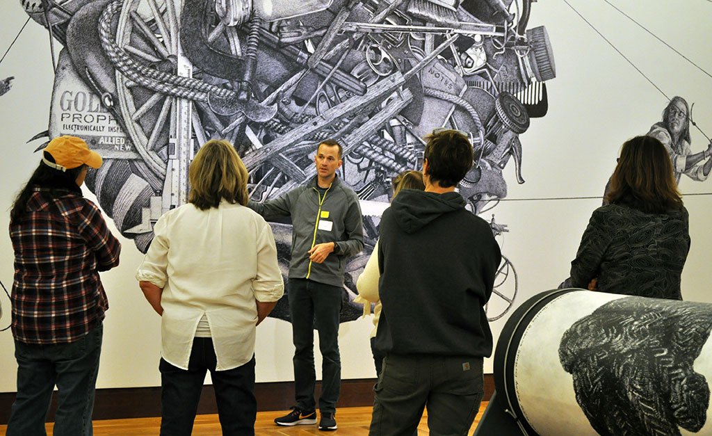 man stands in front of and gestures towards large wall drawing while speaking to a group