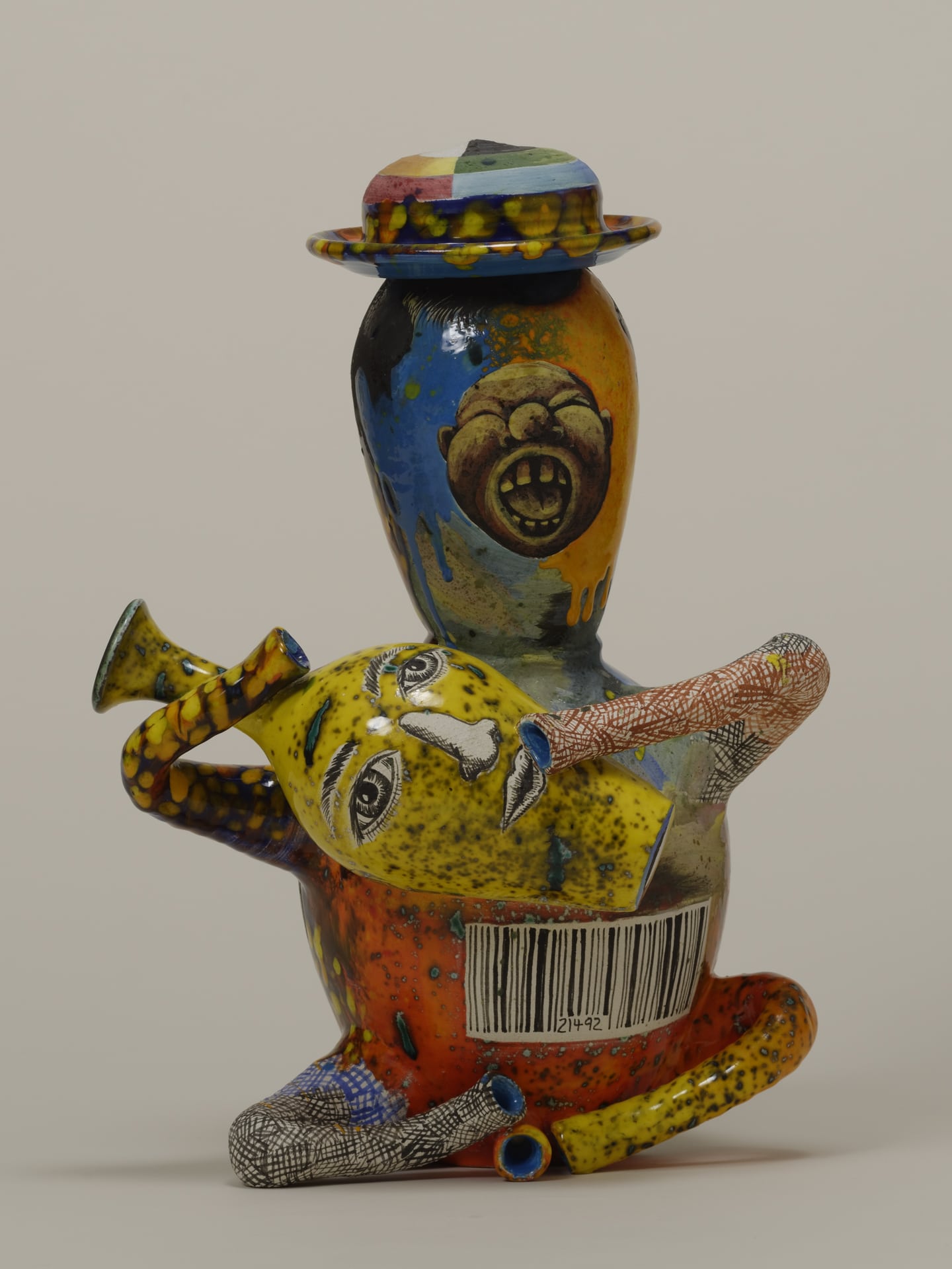 a colorful teapot shaped like a seated man wearing a hat holding a jug with a face on it that forms the spout