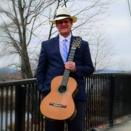 Man in suite and hat holding guitar while standing on a bridge