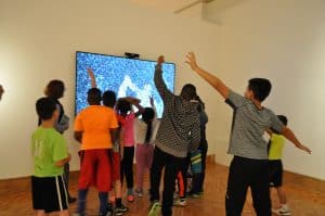 Gorup of youth with backs to  viewer and arms out as they look at work of interactive  art.