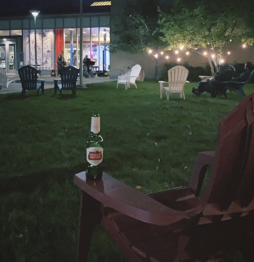 Outside the Currier, it is dark, lights are lit in the trees, and inside the lobby, Adirondack chairs on the lawn, some people sitting there, an empty one in the foreground with a beer bottle on the armrest.