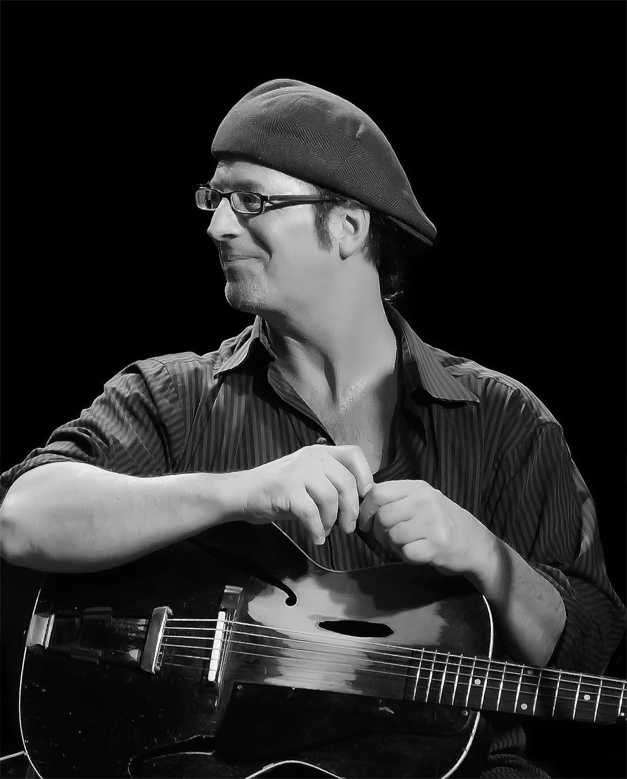 Man smiling to the left, holding a guitar and wearing a cap backwards