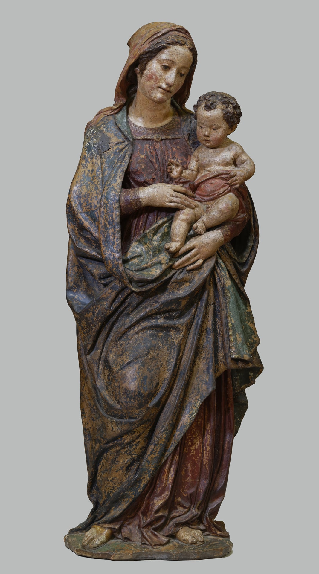 a terracotta statue of the madonna holding the christ child. the paint is worn; she wears a blue robe over a red dress.