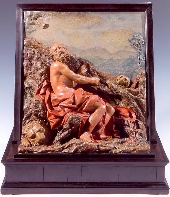 a painted relief of Saint Jerome who wears a cloth over his legs, sitting on rocks gazing up to the sky. a mountainous landscape is in the background; a lion sleeps at his feet.